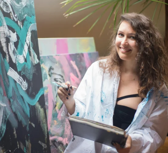 Smiling woman with white man's shirt on sitting in front of an easel brush in hand ready to paint.